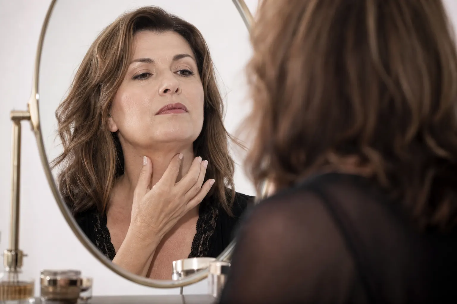 Woman examining her neck skin in a mirror, touching the front of her neck